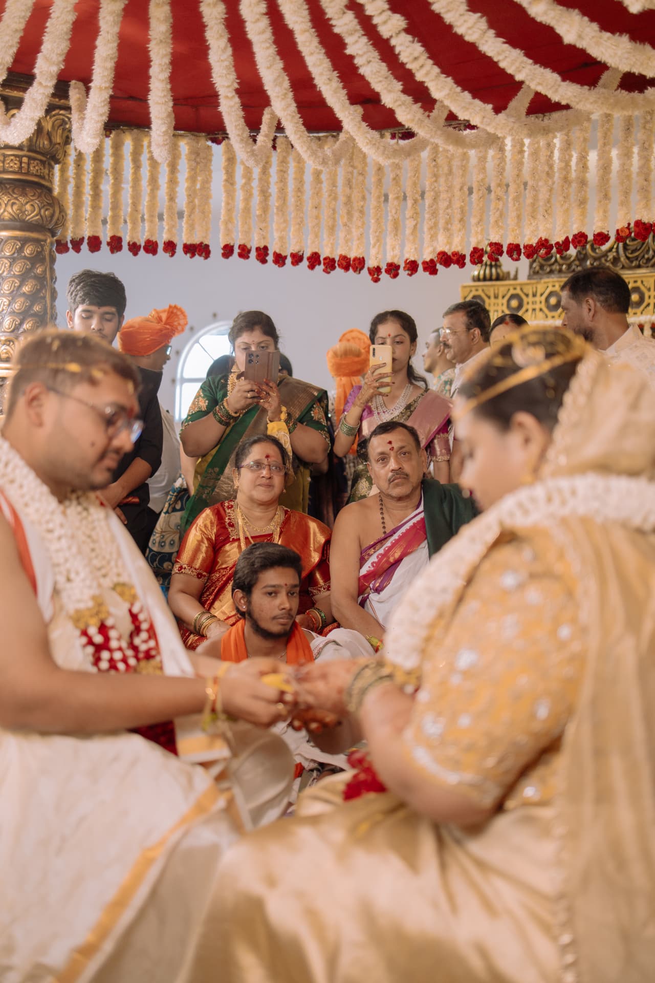 A bride and groom seated beneath a floral wedding canopy, holding hands during a ritual