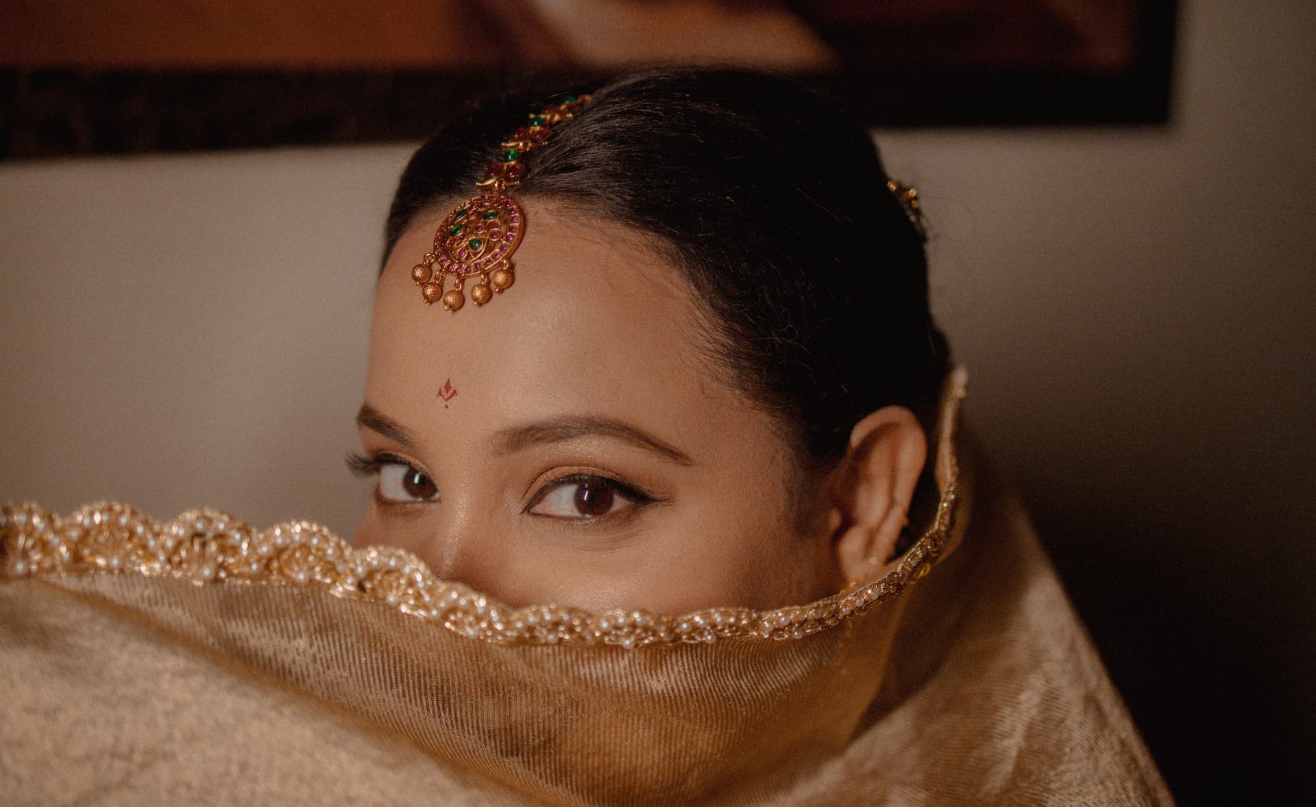 A bride's eyes peeking over a pearl-trimmed veil, wearing a gold maang tikka and jhoomar